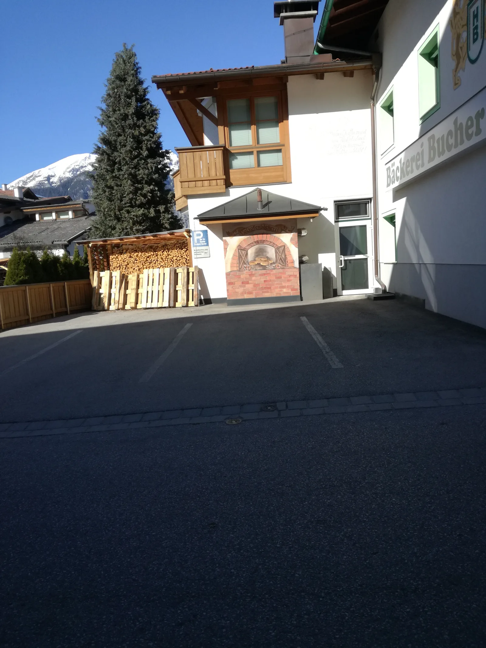 Rückseite der Bäckerei mit dem Wandbild Holzofenbäckerei über dem Ofen, daneben gestapeltes Brennholz